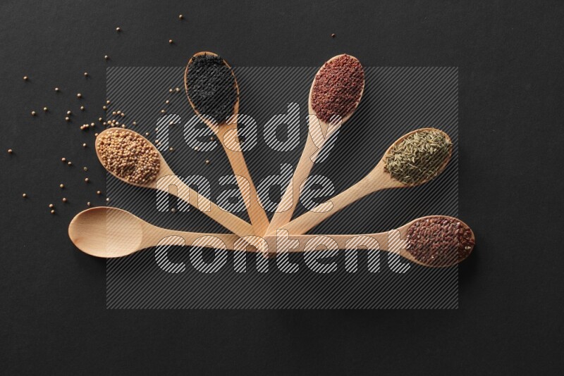 wooden spoons filled with white peppers, mustard seeds, black seeds, garden cress, cumin and flax on black flooring