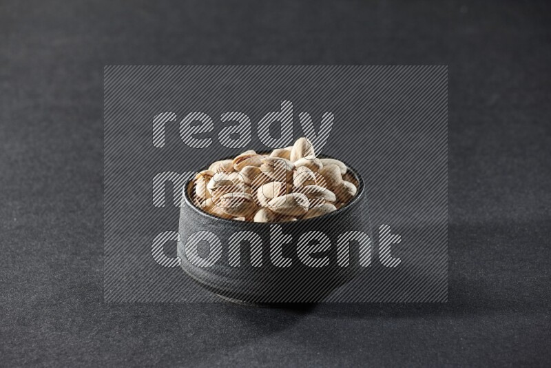 A black pottery bowl full of pistachios on a black background in different angles
