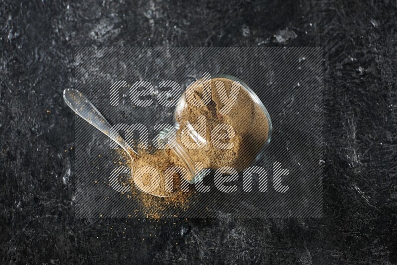 وعاء زجاجي للتوابل مقلوب وملعقة معدنية ممتلئة ببودرة الكمون والبودرة تناثرت منه على خلفية سوداء