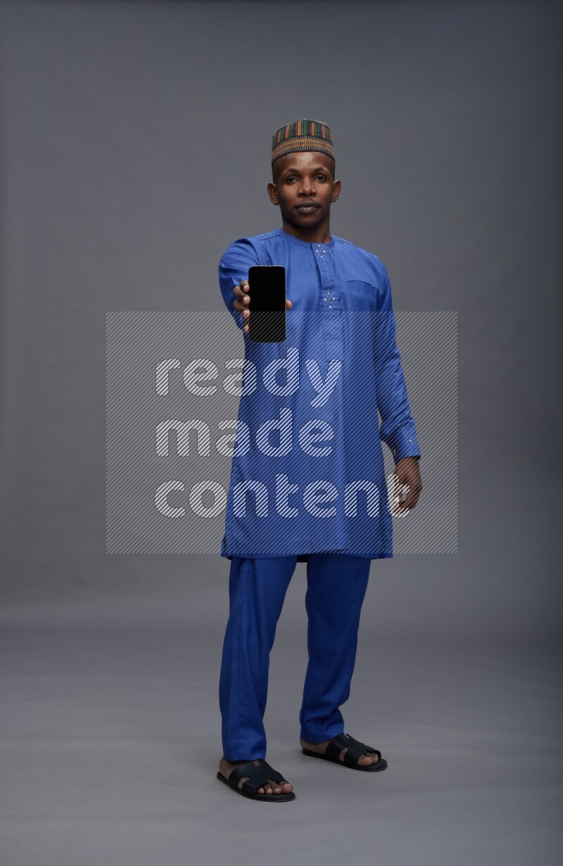 Man wearing Nigerian outfit standing showing phone to camera on gray background