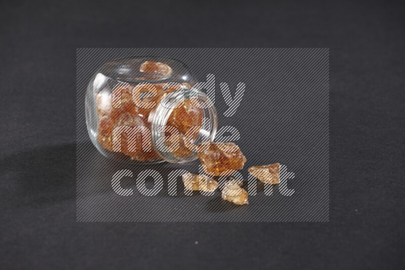 وعاء زجاجي ممتلئ بقطع الصمغ العربي والوعاء مقلوب وبعض القطع متناثرة خارجه علي خلفية سوداء