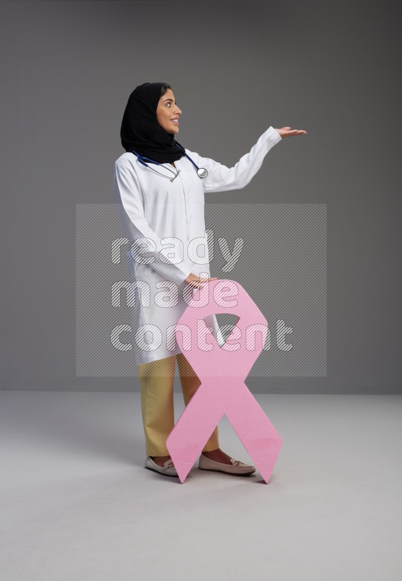 Saudi woman wearing lab coat with stethoscope standing holding awareness ribbon symbol standing on Gray background