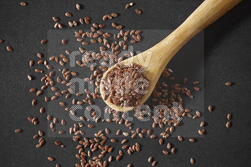 A wooden spoon full of flaxseeds and seeds spread beside it on a black flooring