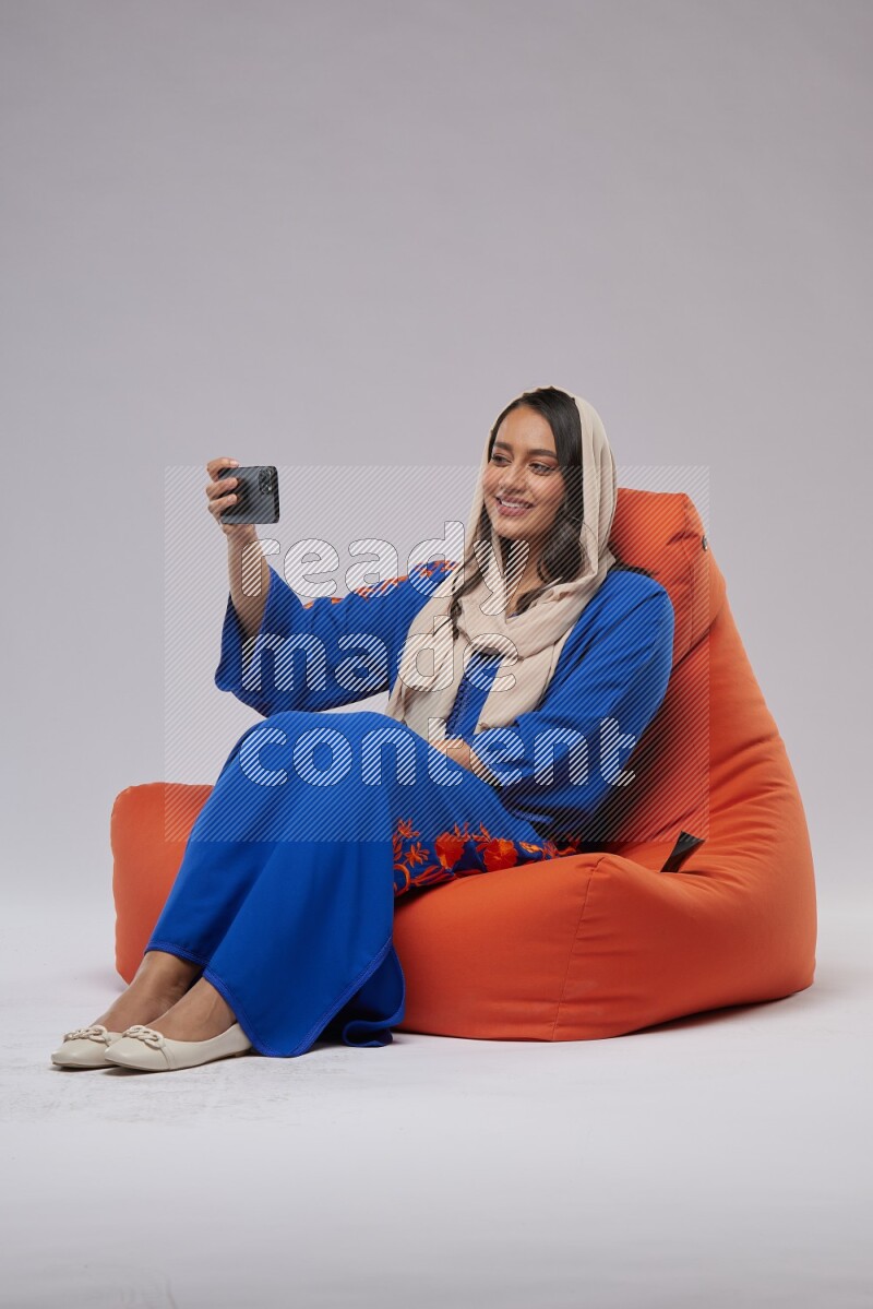 A Saudi woman wearing jalabiya sitting on an orange beanbag and taking selfie