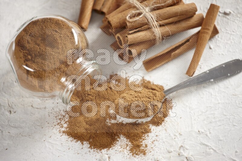 Flipped herbs glass jar full of cinnamon powder with a metal spoon full of powder and cinnamon sticks on a textured white background