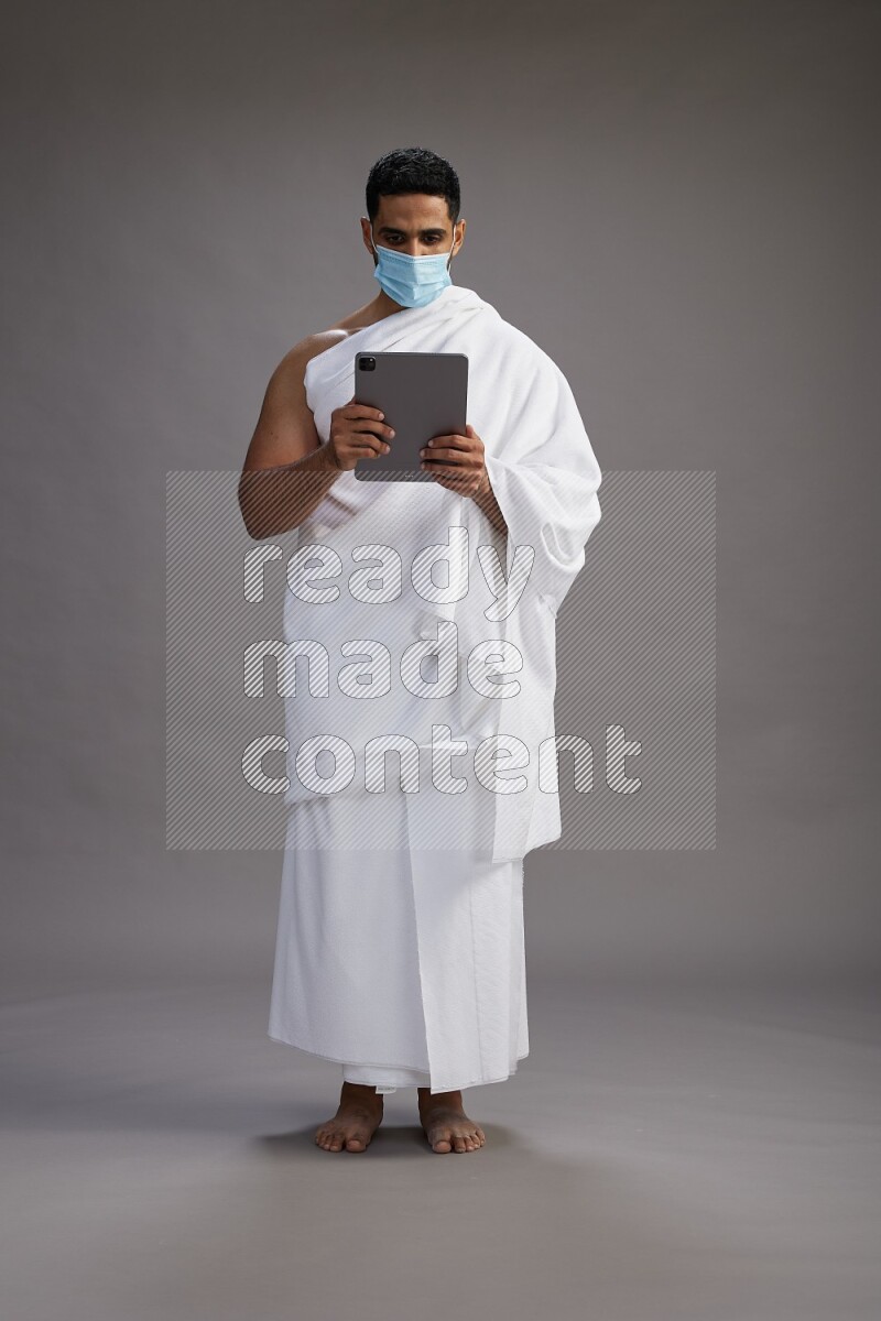 A man wearing Ehram with face mask Standing holding a tablet on gray background