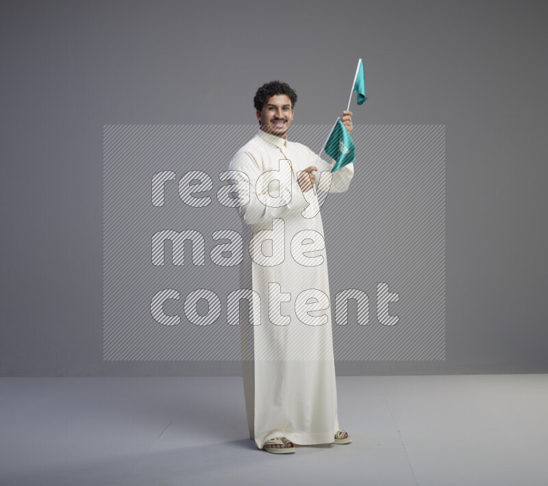 A Saudi man standing wearing thob holding small Saudi flag on gray background