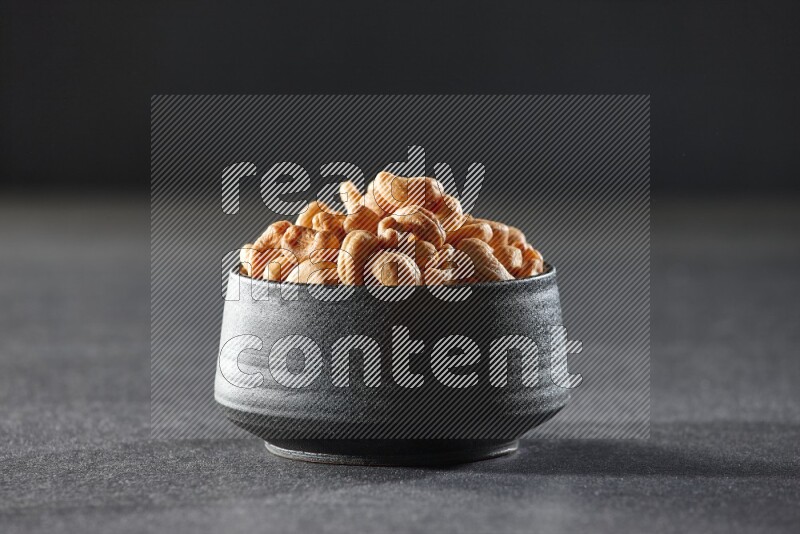 A black pottery bowl full of cashews on a black background in different angles