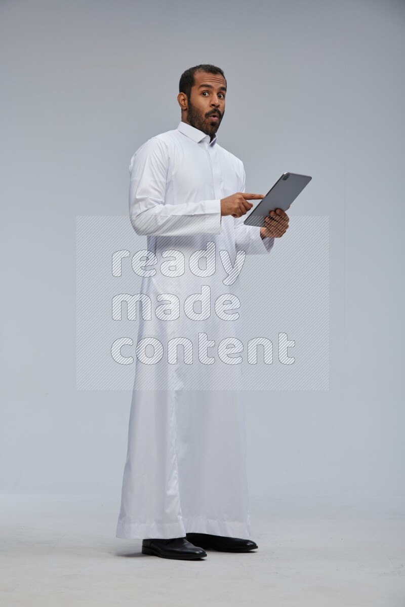 Saudi man wearing Thob standing working on tablet on Gray background