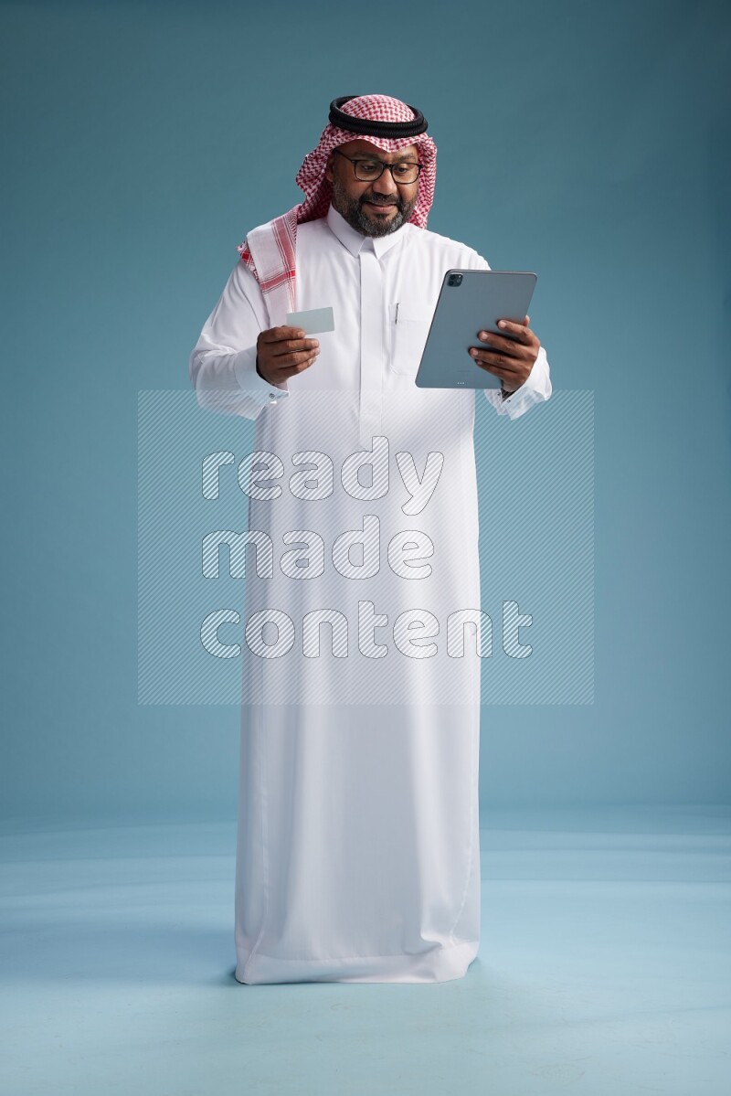 Saudi Man with shimag Standing holding ATM card while working on tablet on blue background