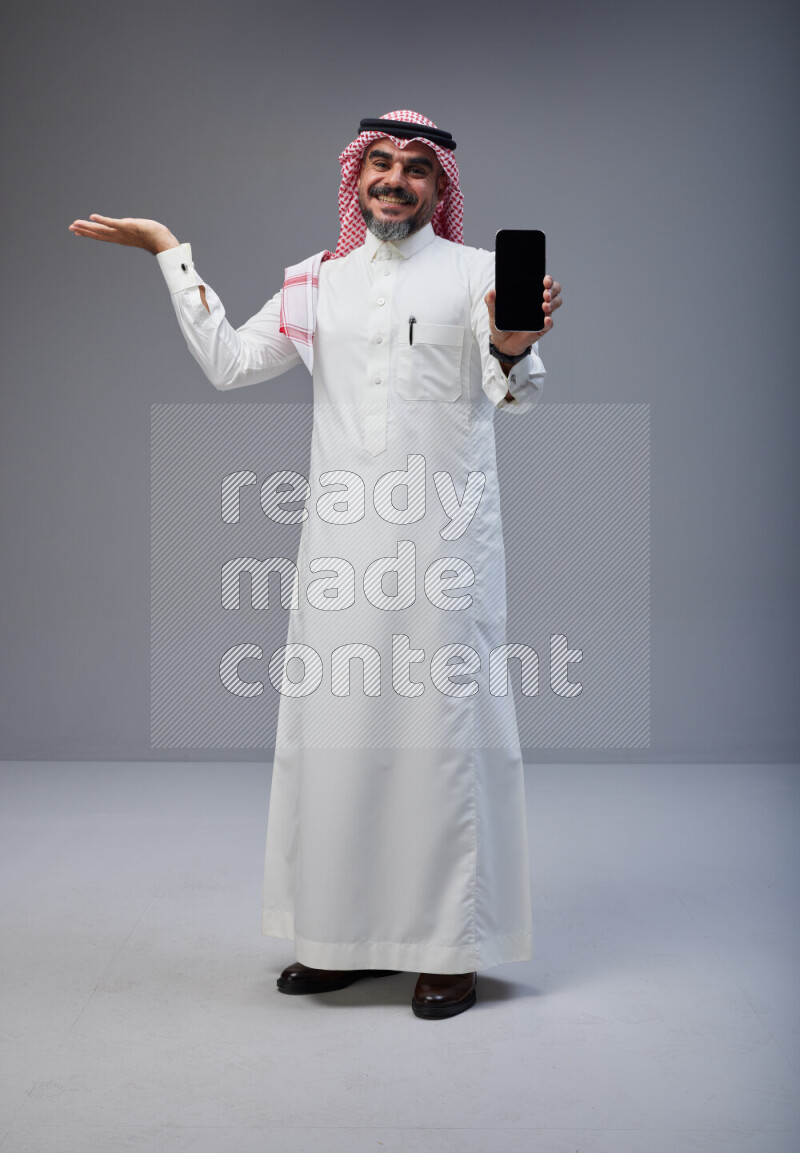 Saudi man Wearing Thob and red Shomag standing showing phone to camera on Gray background
