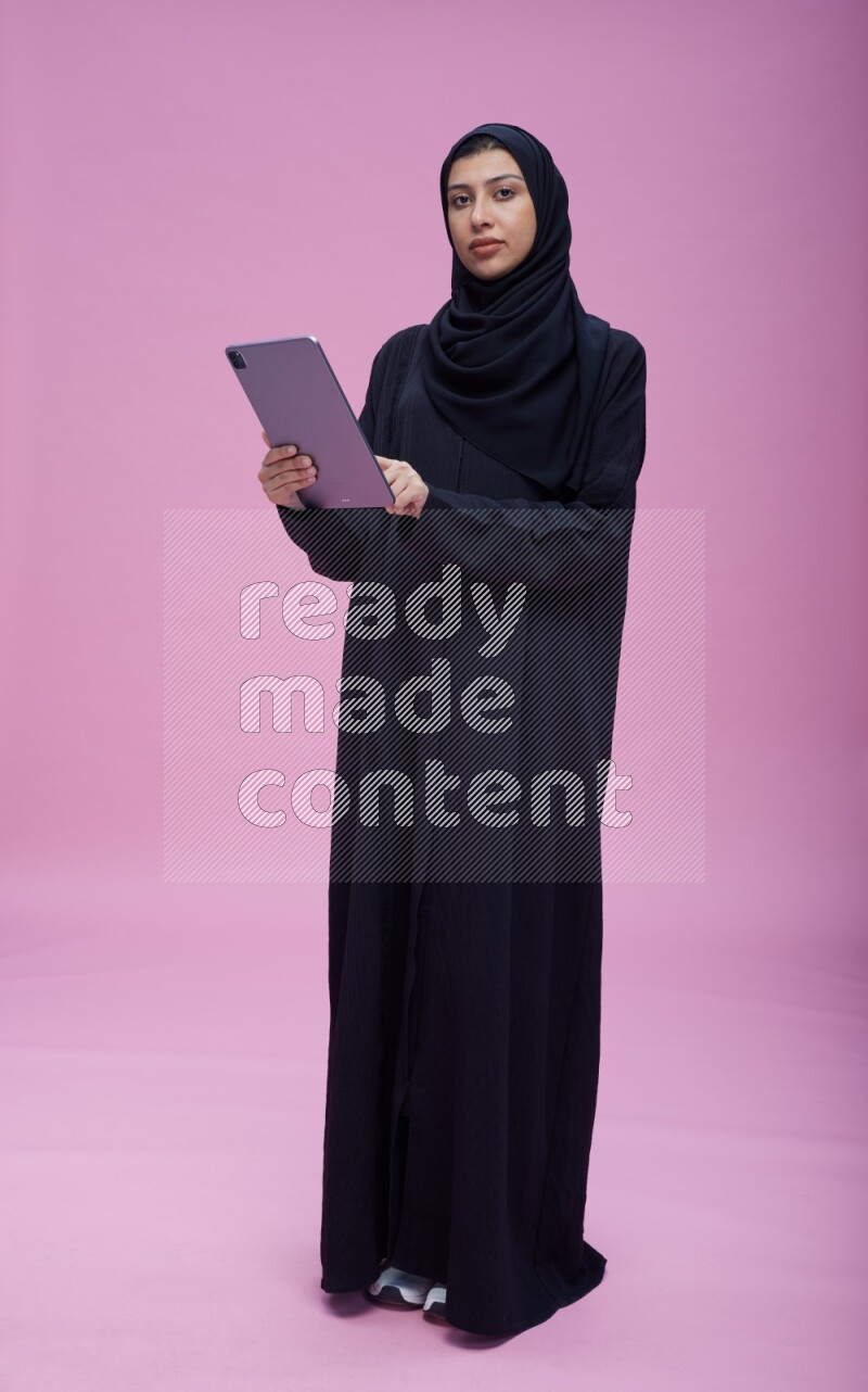 Saudi woman wearing Abaya standing working on tablet on pink background