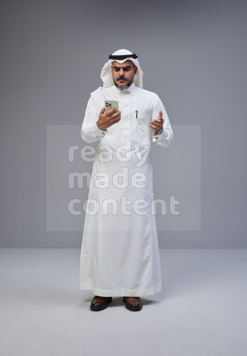 Saudi man Wearing Thob and white Shomag standing texting on phone on Gray background