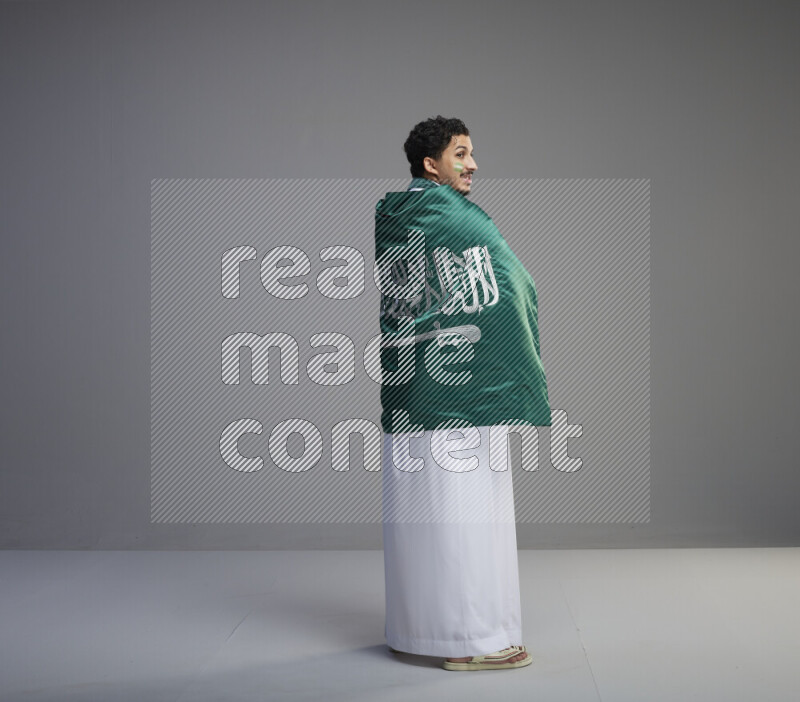 A Saudi man standing wearing thob with face painting wrapping big Saudi flag on gray background