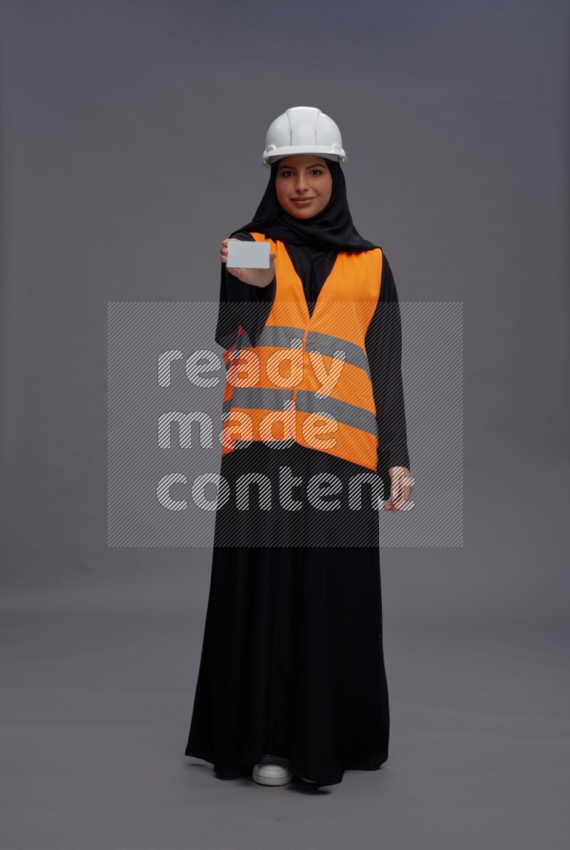 Saudi woman wearing Abaya with engineer vest standing holding ATM card on gray background