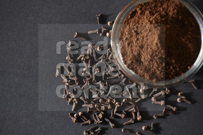 A glass bowl full of cloves powder with cloves grains spread on black flooring