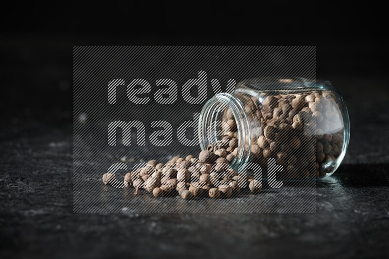 A flipped glass spice jar full of allspice whole balls and the balls spilled out of it on a textured black flooring