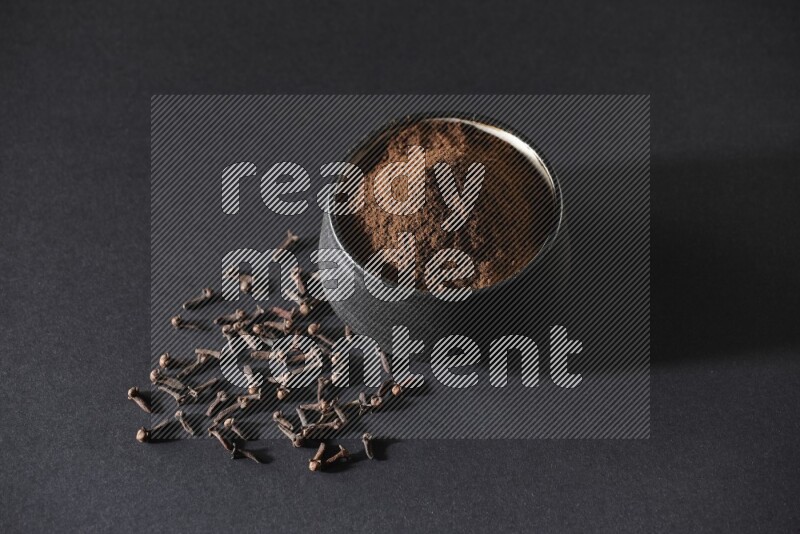 A black pottery bowl full of cloves powder and cloves whole grains on a black flooring