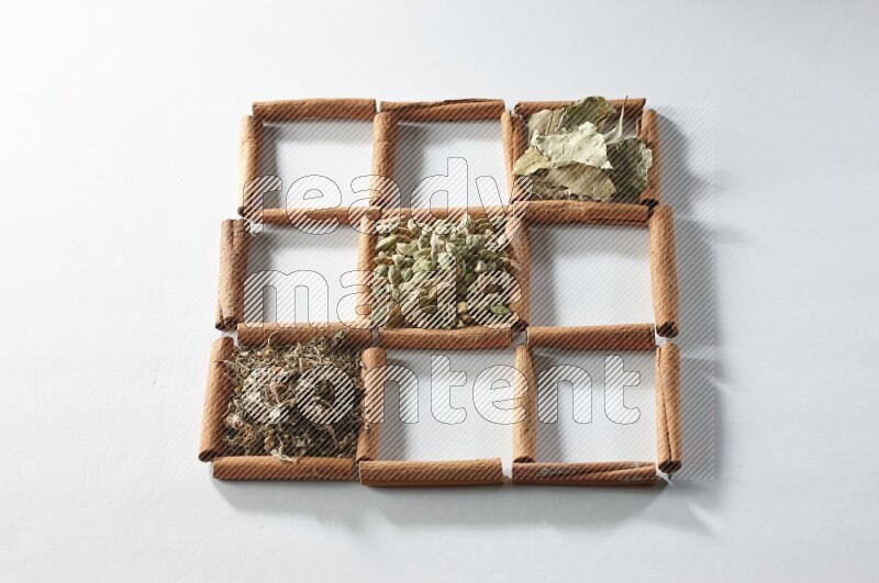 9 squares of cinnamon sticks full of cardamom in the middle surrounded by nutmeg, cinnamon, bay laurel leaves, cloves, cumin, dried ginger, dried basil and star anise on white flooring
