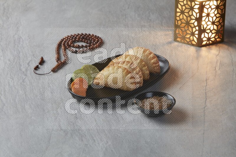 Four fried sambosas in an oval shaped black plate, beside a cut cheese sambosa, a brown misbaha and a golden lantern on a gray background