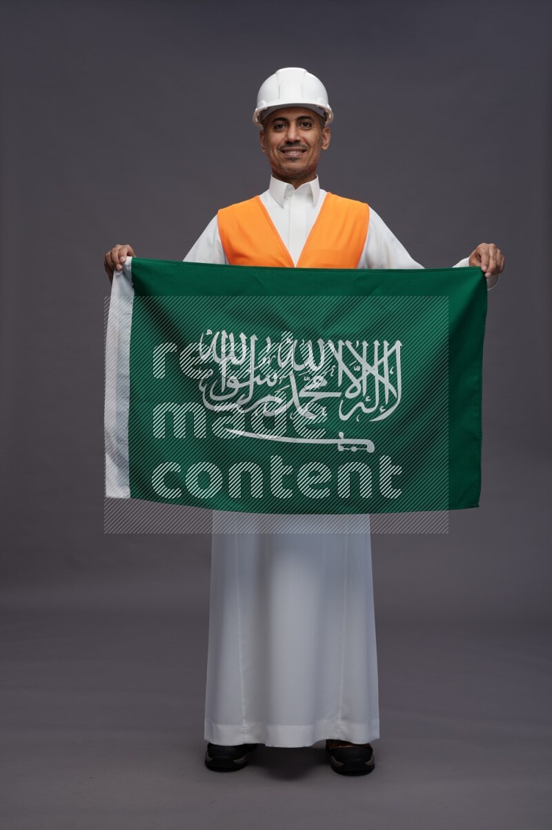 Saudi man wearing thob with engineer vest standing holding Saudi flag on gray background