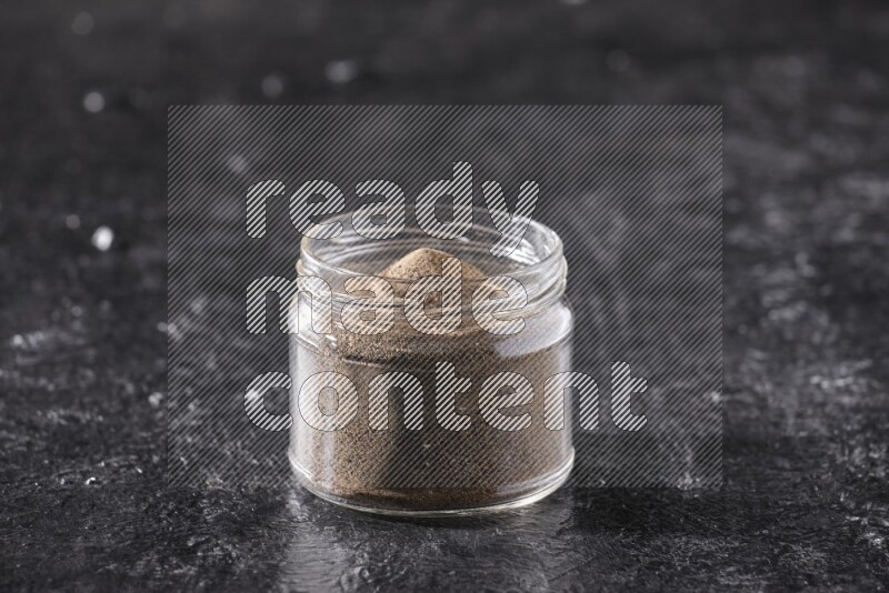 A glass jar full of black pepper powder on a textured black flooring