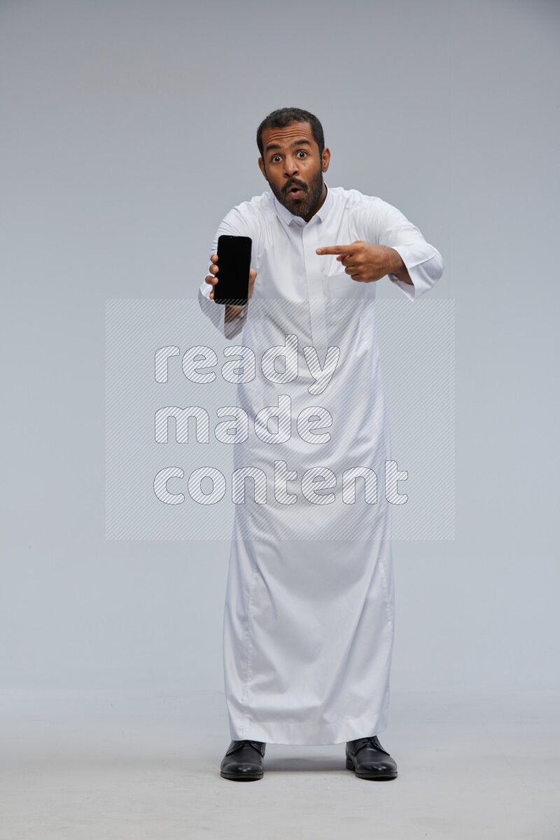 Saudi man wearing Thob standing showing phone to camera on Gray background
