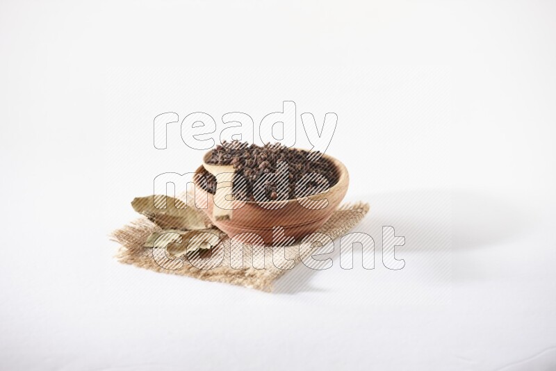 A wooden bowl and a wooden spoon full of cloves on a piece of burlap on a white flooring