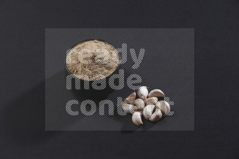 A black pottery bowl full of garlic powder with some cloves beside it on a black flooring