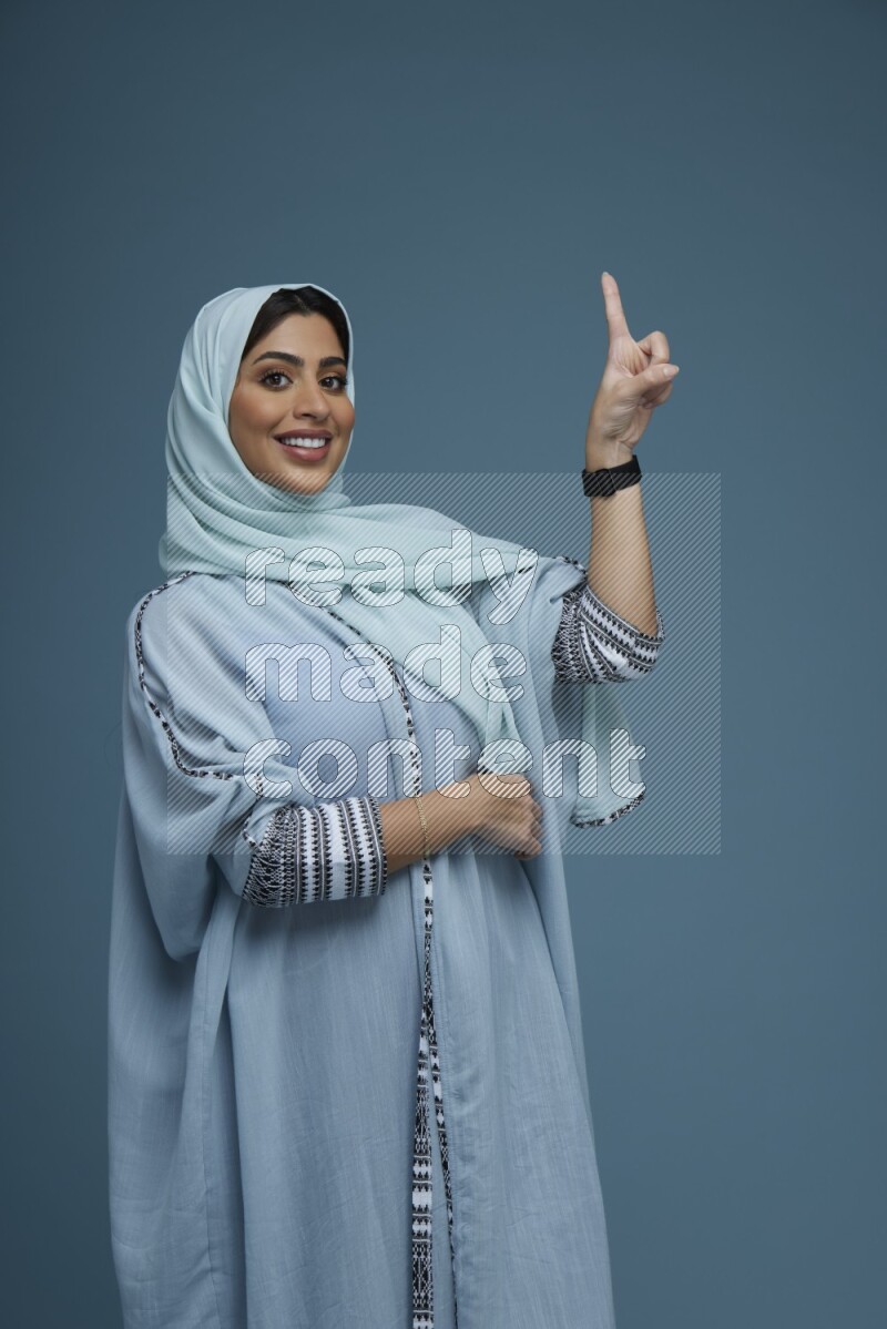 A Saudi woman pointing in a blue background wearing a blue Abaya with hijab