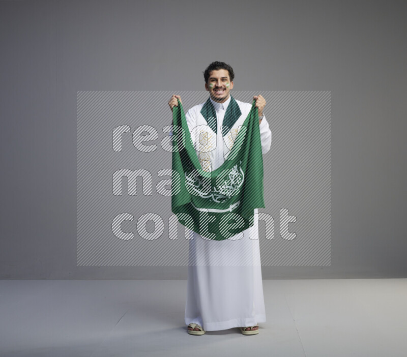 A Saudi man standing wearing thob with face painting wrapping Saudi flag scarf and holding big Saudi flag on gray background