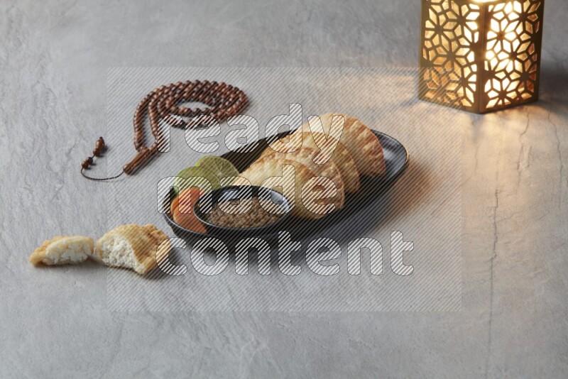 Four fried sambosas in an oval shaped black plate, beside a cut cheese sambosa, a brown misbaha and a golden lantern on a gray background