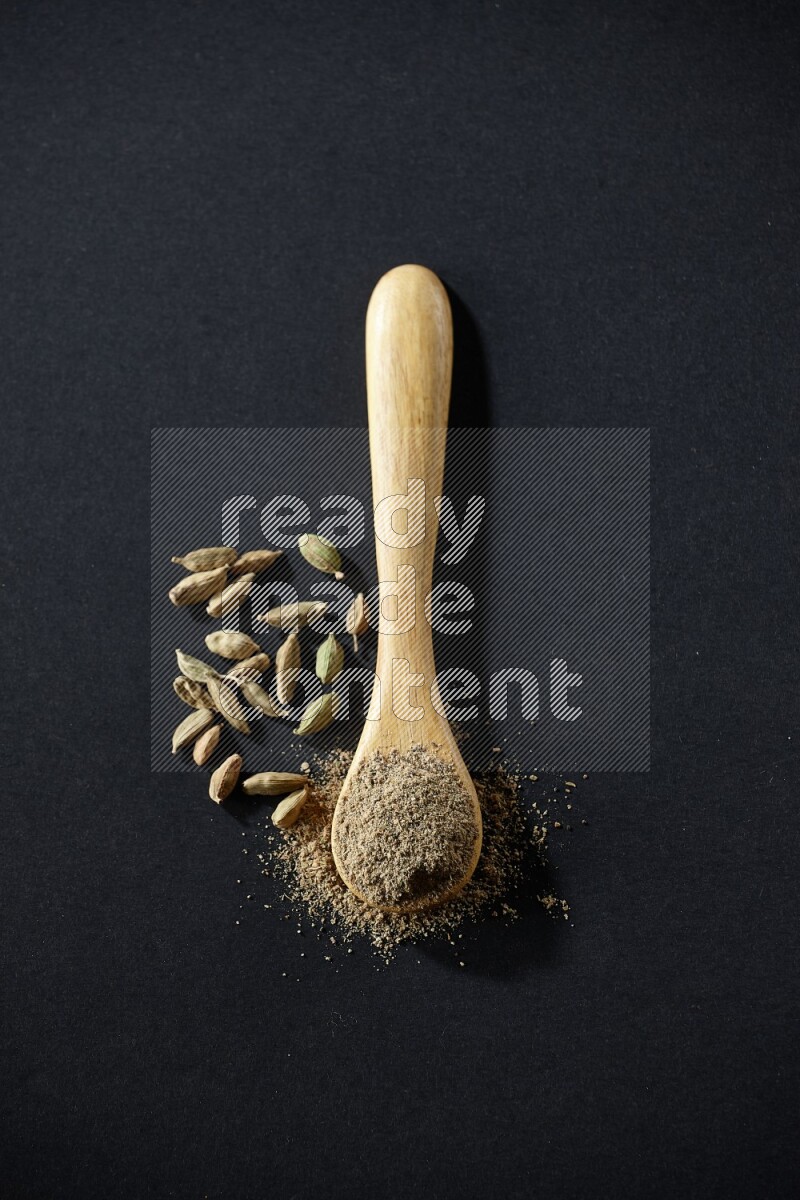A wooden spoon full of cardamom powder and cardamom seeds beside it on black flooring
