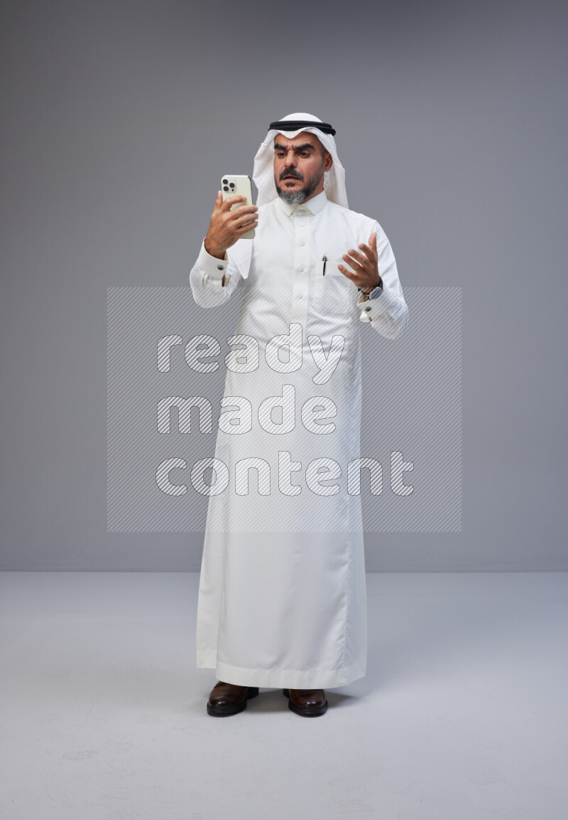 Saudi man Wearing Thob and white Shomag standing texting on phone on Gray background