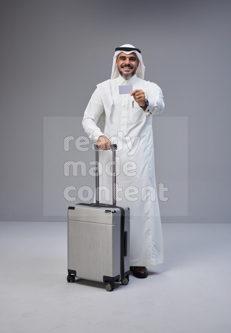 Saudi man wearing Thob and white Shomag standing holding Travel bag and ATM card on Gray background