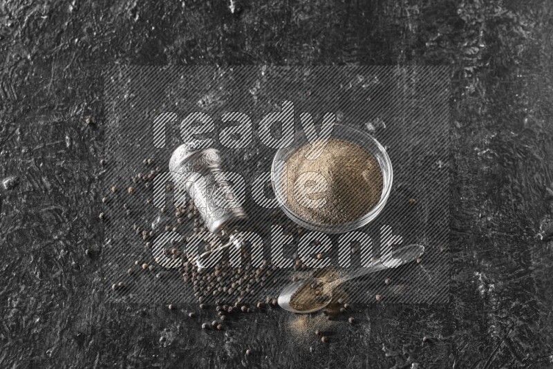 A glass bowl full of black pepper powder with black pepper beads, a turkish metal pepper grinder and a metal spoon on textured black flooring