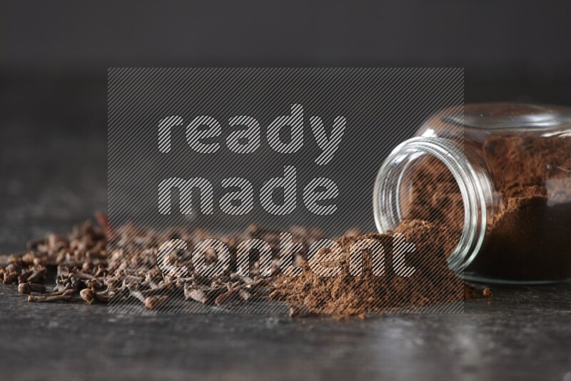 وعاء زجاجي للبهارات مقلوب ممتلئ ببودرة القرنفل والبودرة سقطت منه مع حبوب القرنفل المتناثرة على أرضية سوداء