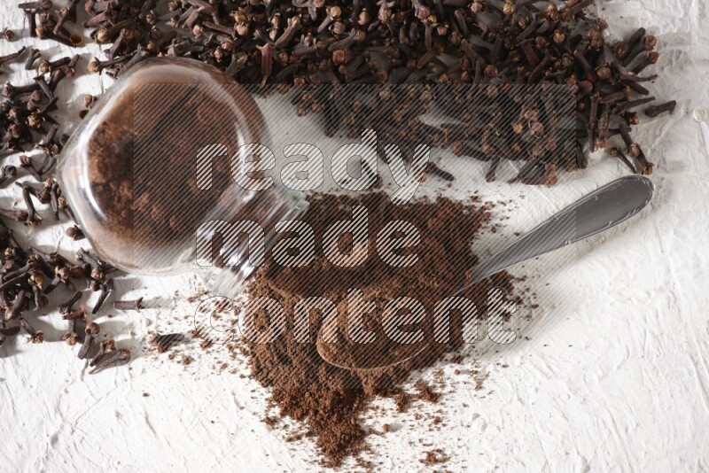 وعاء زجاجي مقلوب ممتلئ ببودرة القرنفل وتناثرت البودرة منه مع حبوب القرنفل المنتشرة على أرضية بيضاء