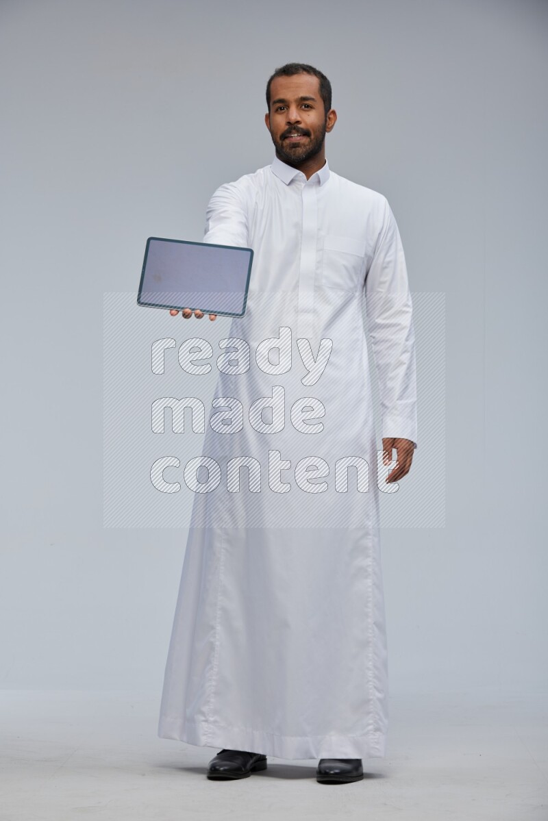 Saudi man wearing Thob standing showing tablet to camera on Gray background