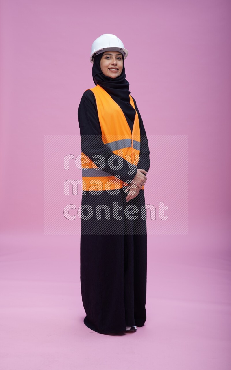 Saudi woman wearing Abaya with engineer vest and helmet standing interacting with the camera on pink background