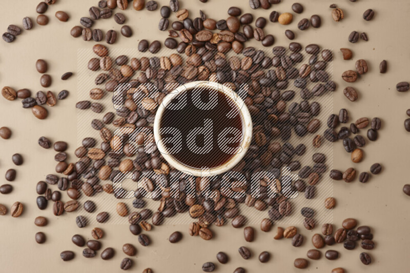 A beige pottery cup of coffee surrounded by roasted coffee beans on beige background