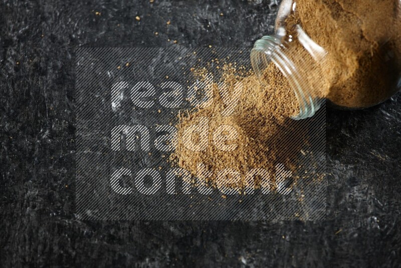 A flipped glass spice jar full of cumin powder and powder spilled out on a textured black flooring