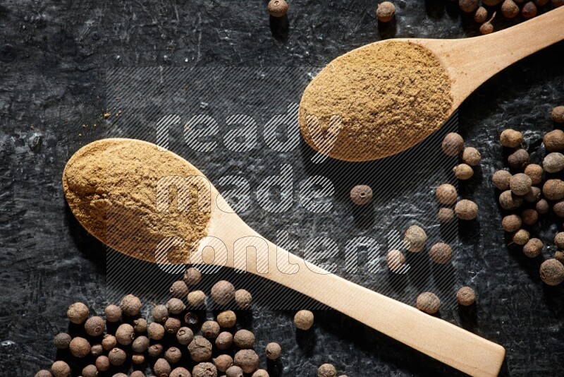 Wooden spoons full of all spice powder and allspice whole balls beside it on a textured black flooring