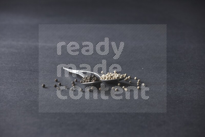 2 metal spoons full of black and white pepper beads with spreaded beads on black flooring