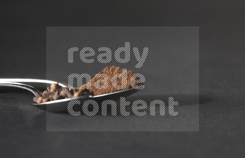 2 metal spoons, one full of cloves grains and the other full of cloves powder on a black flooring
