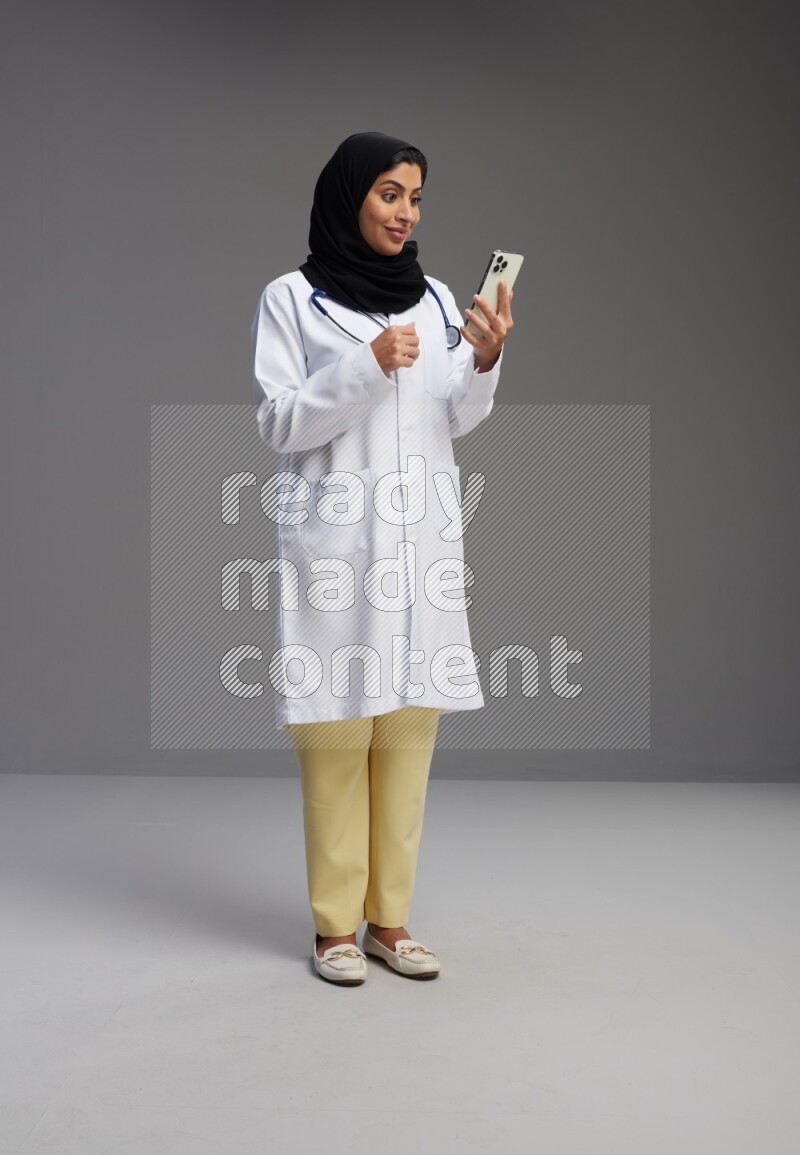 Saudi woman wearing lab coat with stethoscope standing texting on phone on Gray background