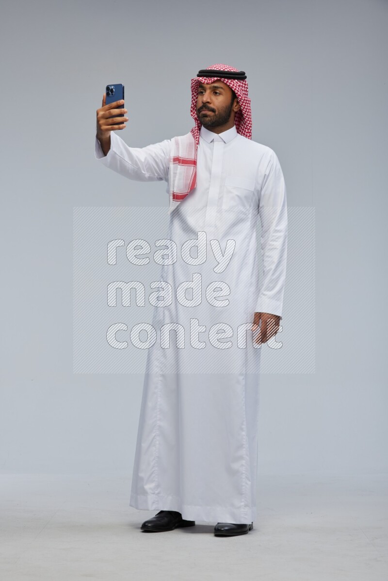 Saudi man Wearing Thob and shomag standing taking selfie on Gray background