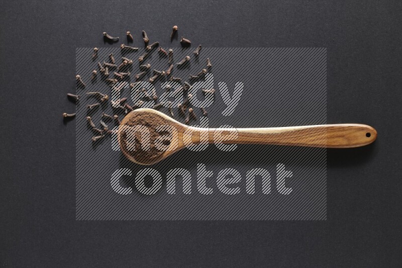 A wooden ladle full of cloves powder and some of whole cloves around it on a black flooring