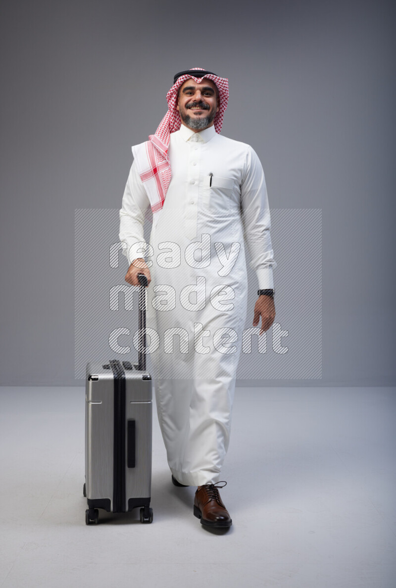 Saudi man wearing Thob and red Shomag standing holding Travel bag on Gray background
