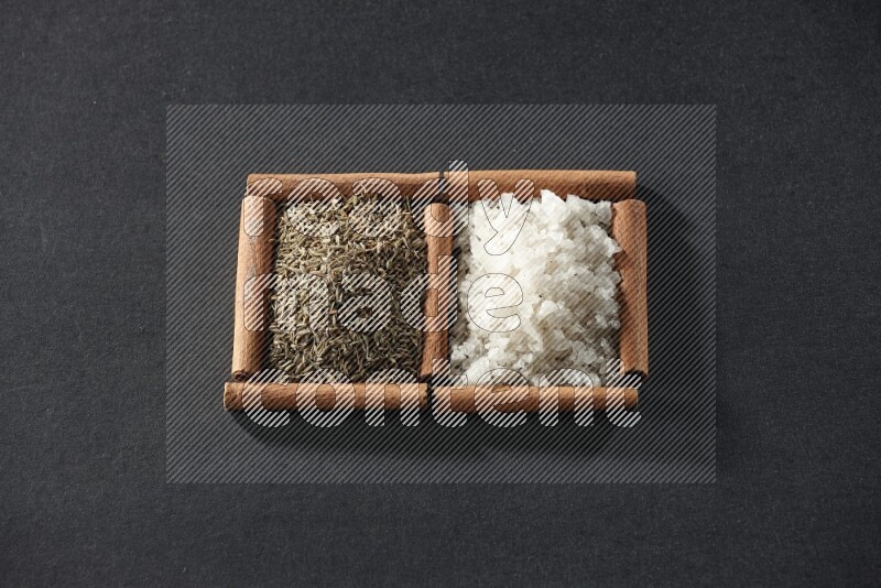 مربعان من أعواد القرفة ممتلئان بالملح الأبيض وحبوب الكمون علي خلفية سوداء